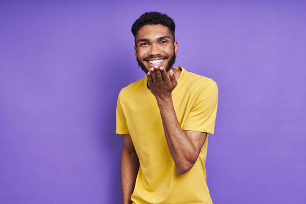 Handsome African man blowing a kiss and smiling while standing against purple background - Фото, изображение