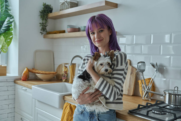 Attractive young woman with purple hair carrying cute little dog while standing at the kitchen - Valokuva, kuva
