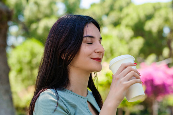 Słodka milenijna kobieta w koszulce stojąca w parku miejskim, pijąca kawę na wynos i patrząca w dal. Poranna koncepcja na świeżym powietrzu. - Zdjęcie, obraz