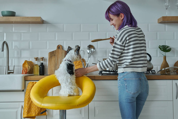 Rear view of young woman cooking at the kitchen while her little dog sitting near her - Foto, Imagem