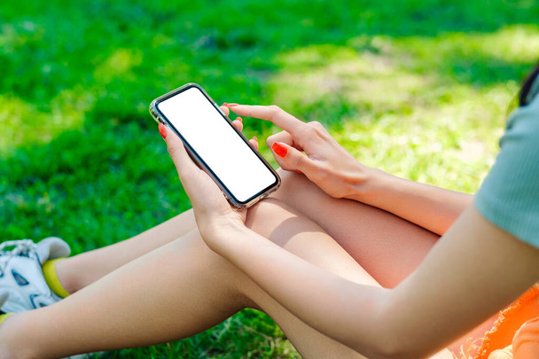 Radosna brunetka w koszulce stojąca w parku miejskim, na zewnątrz ręce trzymające telefon dotykający palca makieta biały pusty wyświetlacz, aplikacja mobilna tech koncepcja, przez ramię widok z bliska. - Zdjęcie, obraz