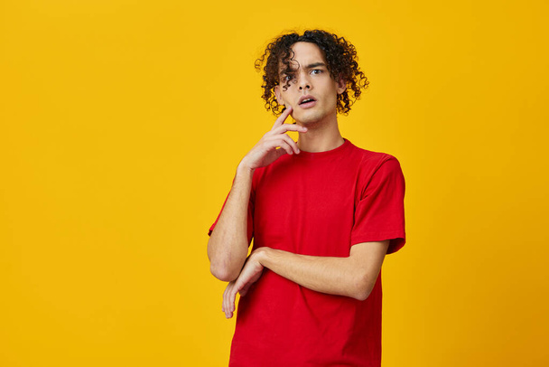Confused funny Caucasian young man in red t-shirt can not make decision posing isolated on over yellow studio background. The best offer with free place for advertising. Emotions for everyday concept - Photo, Image