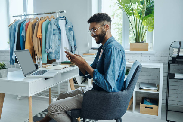 Confident young African man using smart phone while sitting in fashion store office - Photo, Image