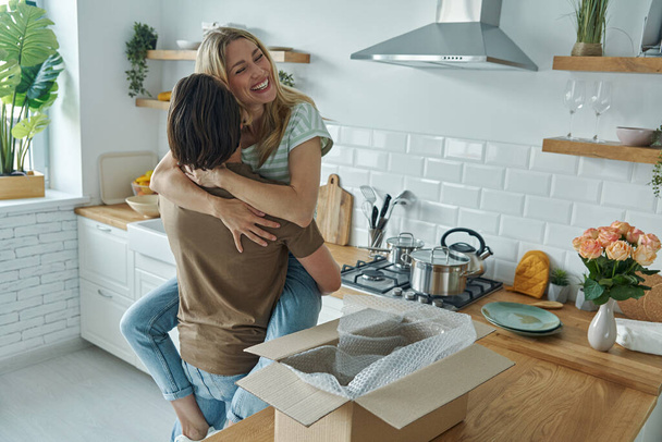 Young couple embracing and smiling while open box laying on the kitchen counter - Foto, Imagen