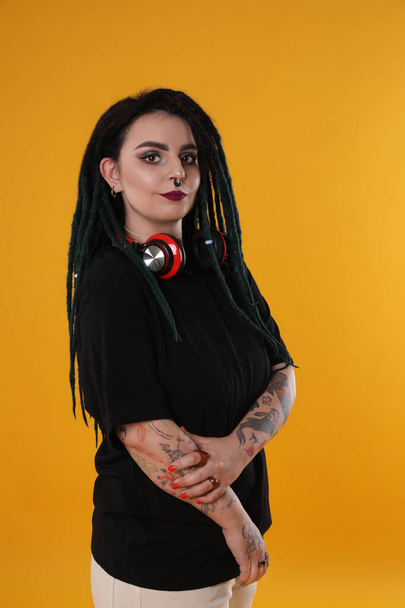 Beautiful young woman with tattoos on arms, nose piercing and dreadlocks against yellow background - Φωτογραφία, εικόνα