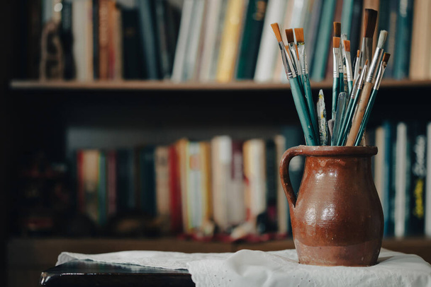 Still life of paint brushes  in clay jug, old books in the background. - 写真・画像