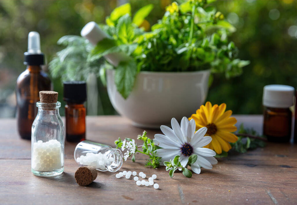 Homeopathy pharmacy, herbal, natural medicine. Homeopathic globules scattered out of a glass bottle, fresh herb in a mortar, green nature background. - Photo, Image