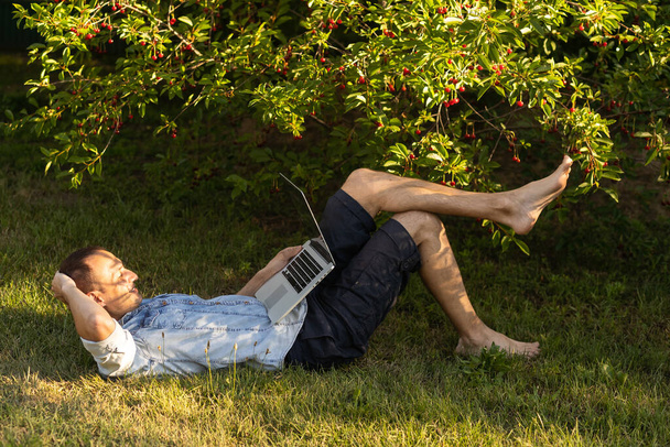 hombre hispano caucásico usando auriculares y sentado en la hierba trabajando con el ordenador portátil. Hombre casual trabajando al aire libre. - Foto, Imagen