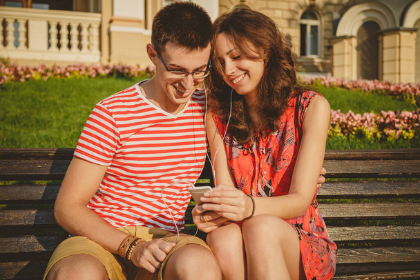 Jonge liefdevolle paar zitten op de bank in het centrum van de stad, lachen en plezier hebben met de smartphone. - Foto, afbeelding