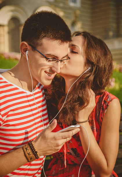 Joven pareja feliz escuchando música y divirtiéndose con el smartphone. Chica está besando a su novio. - Foto, imagen