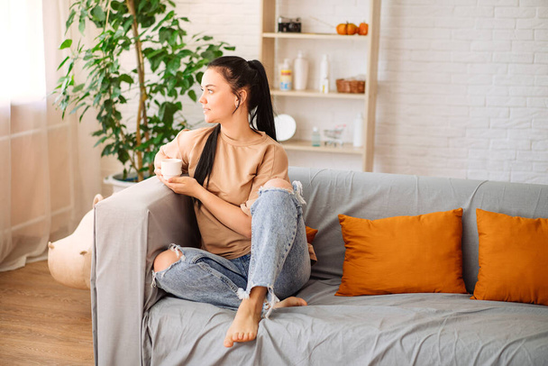 A happy young woman drinks a cup of tea on an autumn morning. A dreamy girl is sitting in the living room with a cup of hot coffee. A beautiful woman at home - 写真・画像