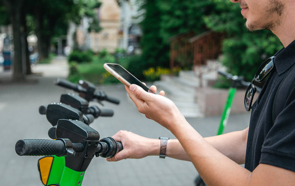 Ηλεκτρικό σκούτερ και κινητό τηλέφωνο. Ένας άνθρωπος που χρησιμοποιεί μια υπηρεσία ενοικίασης με ένα e-scooter σε ένα αστικό δρόμο της πόλης και το πάρκο, χρησιμοποιώντας την ενοικίαση κινητών τηλεφώνων των οικολογικών μεταφορών το καλοκαίρι. - Φωτογραφία, εικόνα