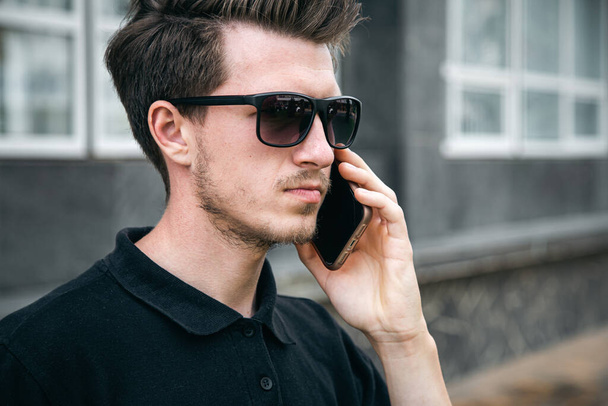 Attractive young man in sunglasses with a cup of coffee talking on a smartphone in the city in the summer on the background of the building. - Foto, afbeelding