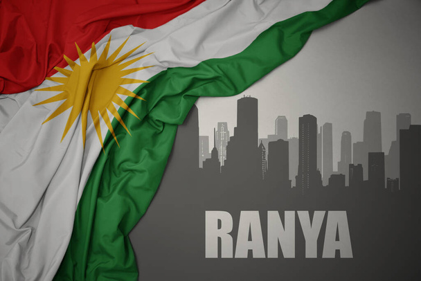 abstract silhouet van de stad met tekst Ranya in de buurt zwaaiende kleurrijke nationale vlag van kurdistan op een grijze achtergrond.3D illustratie - Foto, afbeelding