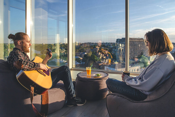 Młody mężczyzna gra na gitarze dla dziewczyny w pobliżu dużego okna z widokiem na miasto, koncepcja daty, przyjaźni i spędzania czasu razem. - Zdjęcie, obraz
