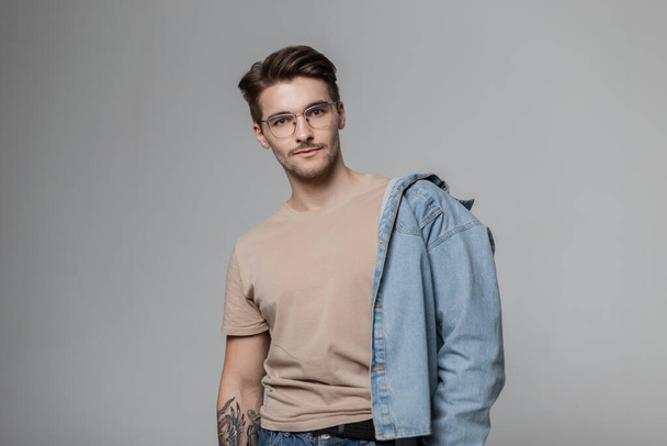ハンサムな男性学生モデルとともに眼鏡とヘアスタイルでファッショナブルなモックアップTシャツとともにジーンズシャツでスタジオで灰色の背景 - 写真・画像
