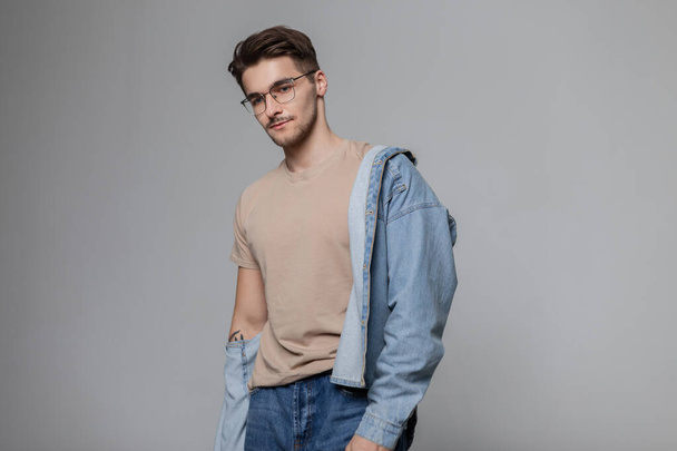 Handsome trendy student guy with hair and vintage glasses in a trendy beige tank top with jeans shirt poses in the studio on a gray background - Fotó, kép