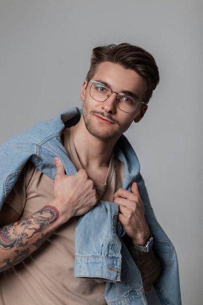 Fashionable handsome model guy with a mustache, beard and haircut in stylish clothes with a T-shirt and tied denim shirt on his shoulders with vintage glasses in the studio, looks at the camera. - Zdjęcie, obraz