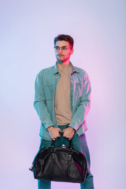 Handsome businessman hipster guy with hairstyle and vintage glasses in casual denim outfit with jeans and leather bag stands in studio with colorful pink and blue lights - Foto, afbeelding