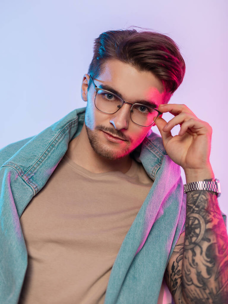 Крутий креативний портрет красивого молодого чоловіка модель з зачіскою в модному джинсовому одязі з татуюваннями на руках носить старовинні модні окуляри в студії з рожевим і синім світлом
 - Фото, зображення