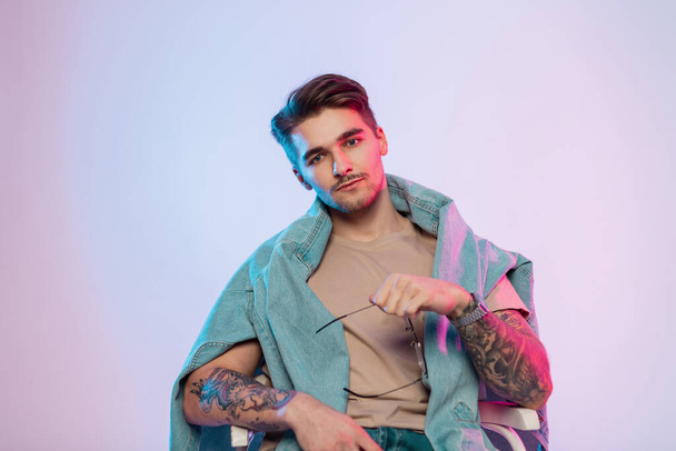 Fashionable cool handsome man model artist with hairstyle and stylish vintage eyewear in casual trendy outfit with tattoos on arms sits in studio with colorful pink blue lights - Foto, imagen