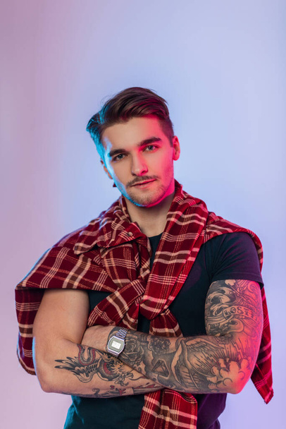 Cool handsome hipster man with tattoos on arms and hairstyle in fashion vintage clothes with plaid shirt in studio with creative pink blue colorful lights - Foto, Imagen