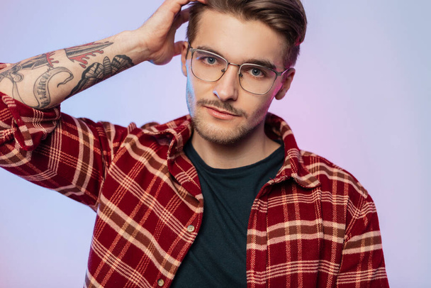 Fashionable handsome hipster guy with hairstyle in vintage stylish outfit with plaid shirt and trendy eyewear in studio with creative colorful lights - Foto, imagen