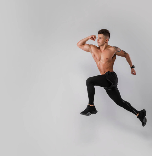 Fashionable handsome young man athlete model with a bare muscular torso in fashionable black sportswear with sneakers jumping and running on a gray background. Copy space - Фото, зображення