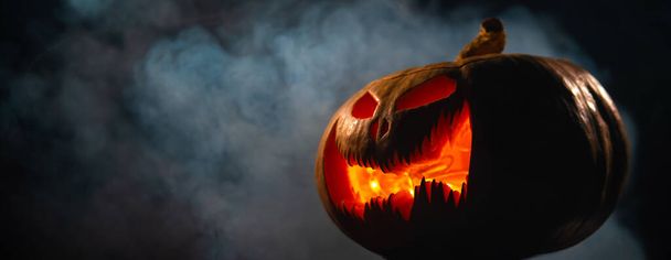 A creepy pumpkin with a carved grimace in the smoke. Jack o lantern in the dark - Фото, изображение