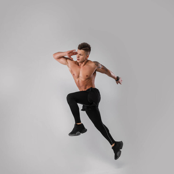 Sporty handsome athlete guy with hairstyle with a naked muscular torso in sportswear with sneakers jumping and running on a gray background. Healthy lifestyle and sports - Photo, image