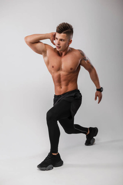 Handsome athletic man fitness model with a bare muscular torso in black sportswear with sneakers doing warm-up and exercise in the gray background - Fotoğraf, Görsel