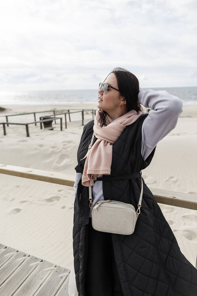 Beautiful young woman with vintage sunglasses in a fashionable black sweatshirt, vest and handbag walks on the beach near the ocean on a spring day - Foto, Imagen