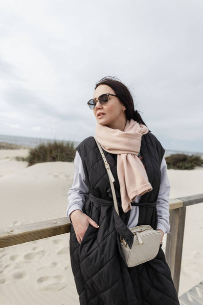 Beautiful fashionable woman with cool vintage sunglasses in a stylish black vest with scarf and sweatshirt walks on the beach by the ocean - Valokuva, kuva