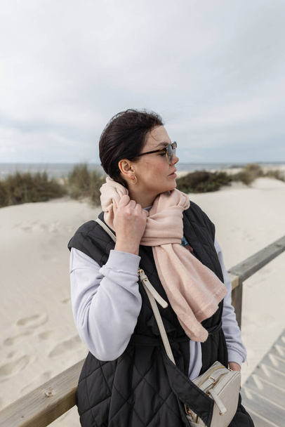 Beautiful young stylish woman with vintage sunglasses wearing fashionable spring outwear with a pink scarf, vest, and pullover bag walks on the beach by the sea - Photo, Image