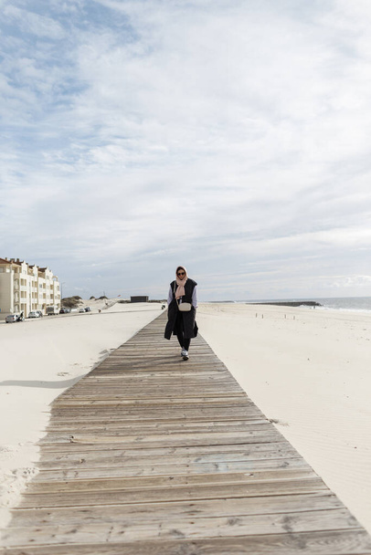 Bella donna alla moda con una sciarpa in testa che indossa un gilet, una felpa e scarpe da ginnastica cammina su una passerella di legno sulla spiaggia nell'oceano - Foto, immagini