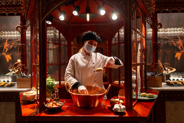 Красивая женщина-повар в форме в медицинской защитной маске готовит блюдо в ресторане. Девушка работает на кухне. Кухня и питание Мастер-класс - Фото, изображение