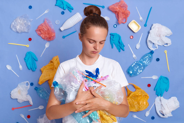 Indoor shot of unhappy sad woman holding collected plastic litter, sad by pollute environment where we live, wearing casual clothes, looking with upset expression at garbage, stands against blue wall. - Foto, imagen