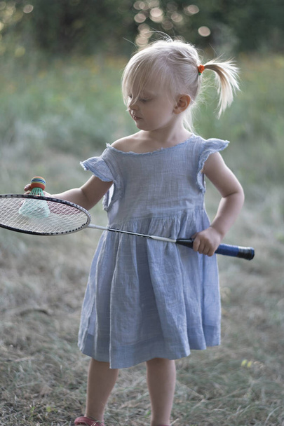 Three-year-old child plays badminton. Cute little girl in summer blue dress holds badminton racket. Vertical frame. - Фото, изображение