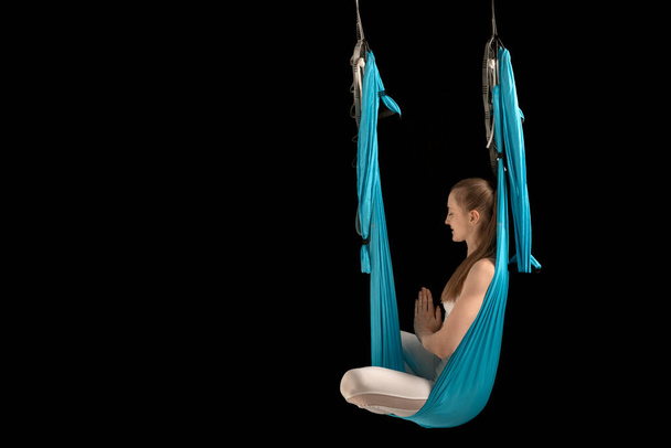 Young woman in lotus pose sits in hammock. Practicing aerial yoga, black background. Copy space. Anti-gravity yoga. - Photo, Image