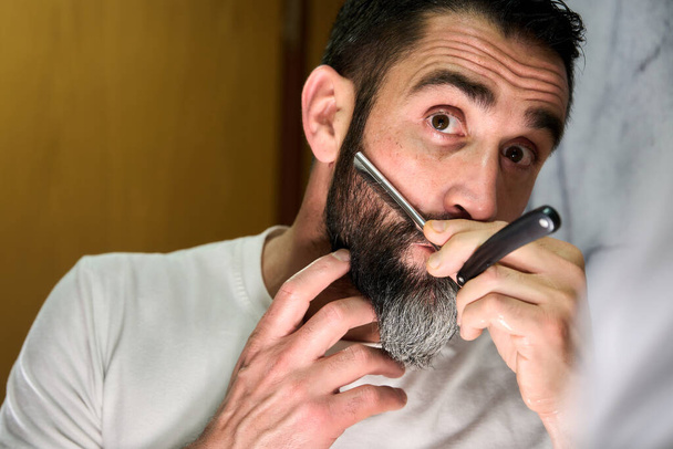 Young man with a beard shaving with a traditional knife in front of the mirror. High quality photo - 写真・画像