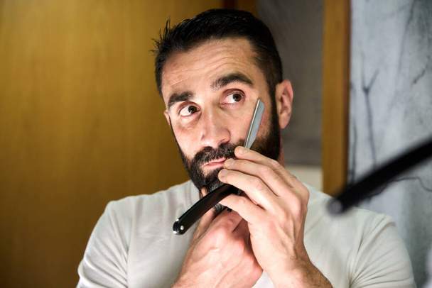 Egy szakállas fiatalember borotválkozik egy hagyományos késsel a tükör előtt. Kiváló minőségű fénykép - Fotó, kép
