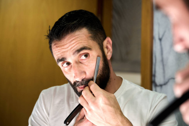 Jovem de barba barbeada com uma faca tradicional à frente do espelho. Foto de alta qualidade - Foto, Imagem