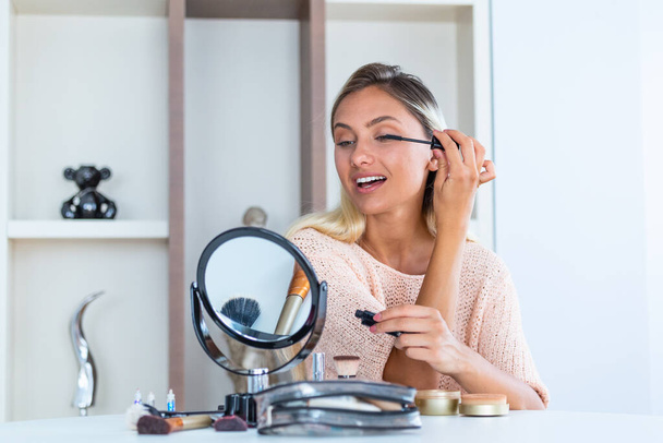 Beautiful woman applying make up (paint her eyelashes) for a evening date in front of a mirror. Portrait Of Beautiful Young Woman Applying Black Mascara On Lashes, Holding Brush In Hand. - 写真・画像