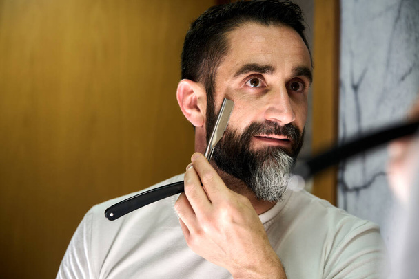 Jeune homme avec une barbe rasée avec un couteau traditionnel devant le miroir. Photo de haute qualité - Photo, image