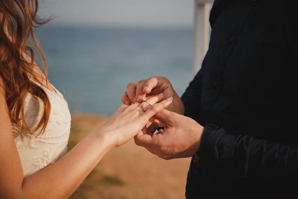 Мужчина надевает обручальное кольцо на руку невесты, закрывается. Свадебная церемония на открытом пляже, стильный жених и невеста стоят возле свадебного алтаря на берегу моря. - Фото, изображение