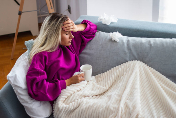 Sick woman with headache sitting under the blanket. Sick woman with seasonal infections, flu, allergy lying in bed. Sick woman covered with a blanket lying in bed with high fever and a flu, resting. - 写真・画像