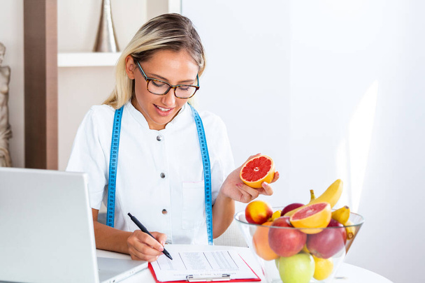 Retrato de una joven nutricionista sonriente en la sala de consulta. Escritorio nutricionista con fruta sana, jugo y cinta métrica. Dietista trabajando en plan de dieta
. - Foto, Imagen