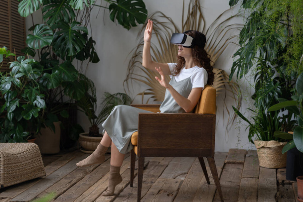 Fiatal lenyűgözött nő visel VR sisak ül karosszékben az üvegházban, monstera deliciosa növény a háttérben. Spanyol nő megtapasztalja a virtuális valóság, miközben pihen a gyönyörű beltéri kertben - Fotó, kép