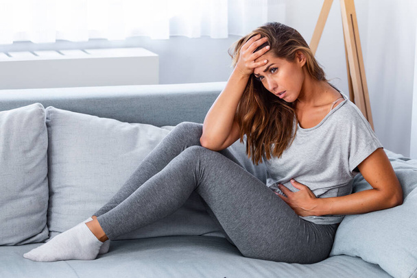View of young woman suffering from stomachache on sofa at home. Woman sitting on bed and having stomach ache. Young woman suffering from abdominal pain while sitting on sofa at home - Photo, Image