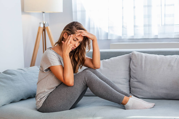 Portrait of an attractive woman sitting on a sofa at home with a headache, feeling pain and with an expression of being unwell. Upset depressed woman lying on couch feeling strong headache migraine. - Фото, изображение